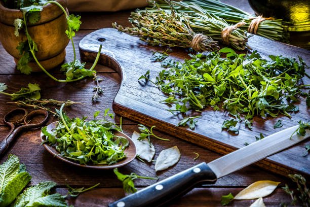 Cutting board with chopped parsley and a kitchen knife on top surrounded by a wooden tray with more chopped parsley, some dried bay leaves and at the background are a few aromatic herbs like thyme, chive, and rosemary tied in bunches. At the left top corner is a mortar with coriander and a vintage scissor. Low key DSLR photo taken with Canon EOS 6D Mark II and Canon EF 24-105 mm f/4L