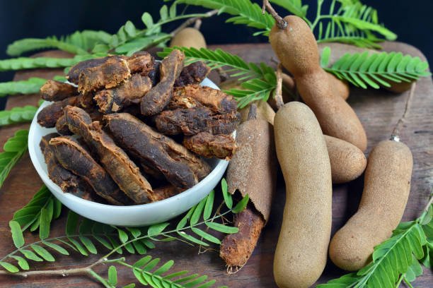 Tamarind paste or juice in white bowl on the wooden table and beside the ripe tamarind and tamarind leaves scattered with black background